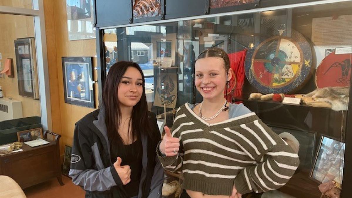 Two young women giving thumbs up while standing in front of an Indigenous art display in their high school foyer