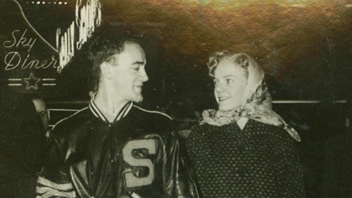 A black and white photo from the 1950s of a man in a letter jacket and a women in a long coat and kerchief smiling at one another and walking at night while holding hands