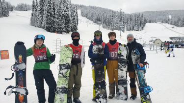 Four smiling teenaged students and their teacher/coach in winter clothing standing in front of  chairlift at ski resort leaning on their snowboards.