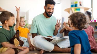 A man reads a book to young children while they are all seated cross-legged on the floor.