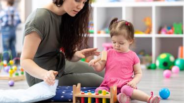 Woman with brown hair plays with a little girl in a pink dress on the floor of a daycare