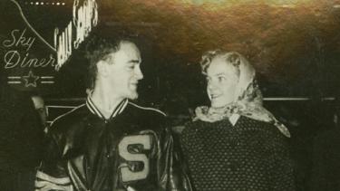 A black and white photo from the 1950s of a man in a letter jacket and a women in a long coat and kerchief smiling at one another and walking at night while holding hands