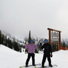 Two snowboarders in front of a ski hill sign at a ski resort