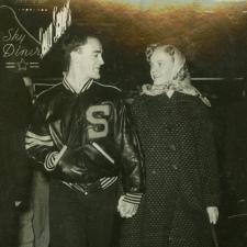 A black and white photo from the 1950s of a man in a letter jacket and a women in a long coat and kerchief smiling at one another and walking at night while holding hands